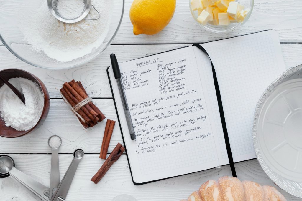 Handskrivet recept i en anteckningsbok bland måttskedar, mjöl och kanel
